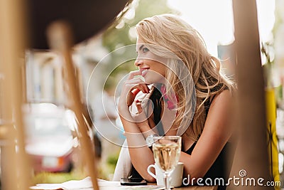 Smiling blonde girl sitting on date at the table with cup of coffee and glass of wine on it. Outdoor blur portrait of Stock Photo