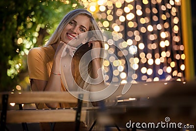 Smiling blonde young woman sitting, with evening fairy lights on the background. Stock Photo