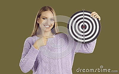 Smiling blondy woman holding dartboard shooting target and pointing finger on khaki background. Stock Photo