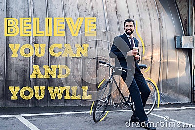 smiling businessman with disposable cup of coffee leaning on bicycle on street with believe you can Stock Photo