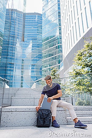 Male freelancer putting laptop inside backpack while sitting on steps on skyscers background Stock Photo