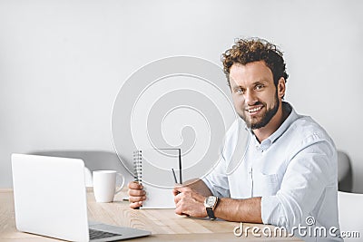 smiling businessman sitting at workplace with laptop and pointing at blank notebook Stock Photo