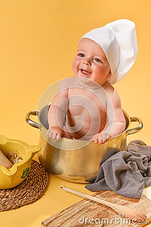 Smiling Caucasian baby tucked inside a pot with chef& x27;s hat and cooking utensils on yellow background Stock Photo