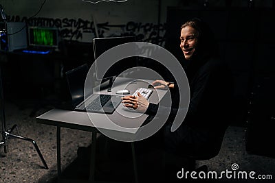 Smiling caucasian hacker man in hood breaking into corporate data servers using laptop and computers sitting at desk in Editorial Stock Photo
