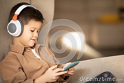 Smiling caucasian little kid in casual, wireless headphones watch video on phone sits in armchair in living room Stock Photo