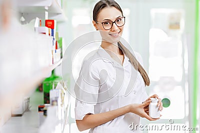 A smiling charming slim dark-haired lady with glasses, wearing a white coat, stands next to the shelf and shows a small Stock Photo