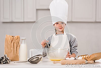 smiling child in chef hat whisking eggs in bowl at table Stock Photo