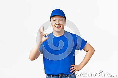 Smiling confident asian courier in blue uniform, wink and showing okay gesture. Delivery guy say OK, guarantee safety Stock Photo