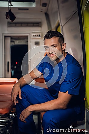 Smiling corpsman in uniform sits and squints in the ambulance car beside stretcher Stock Photo