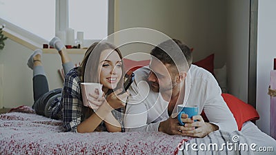 Smiling couple watching TV while lying in bed and drinking coffee at home in the morning Stock Photo