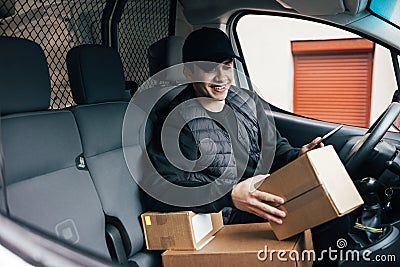 Smiling courier sitting on a driver`s seat checking delivery information on mobile phone while holding box Stock Photo
