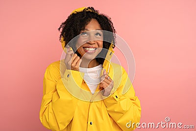 Smiling dark skinned curly woman in yellow raincoat with hood has good mood, looking up at rain and blank space for advertising, Stock Photo