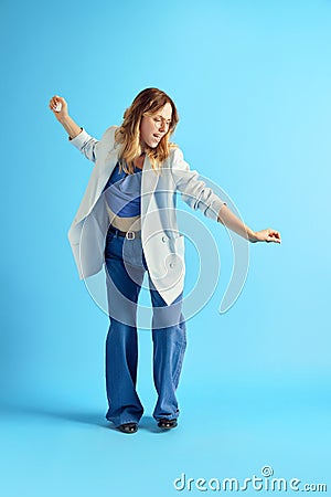 Smiling, delightful young woman listening to music in earphones, dancing and singing against blue studio background Stock Photo