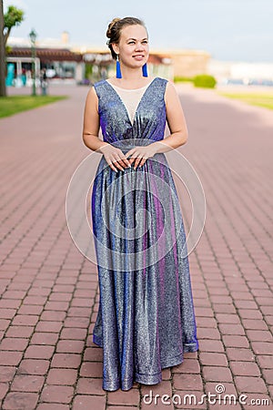 Smiling dreamy young Caucasian woman in amethystine floor-length sparkly dress on pavement Stock Photo