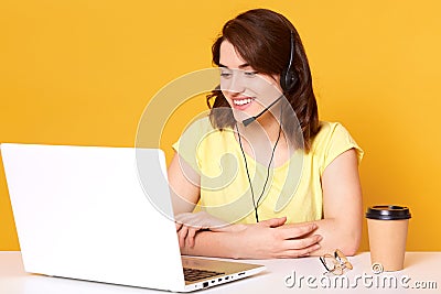 Smiling energetic female sitting at office desk with folded arms, having headset, looking at laptop screen, papercup of coffee and Stock Photo