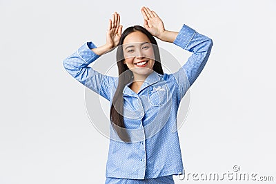 Smiling enthusiastic cute asian girl in blue pajamas showing bunny ears gesture over head, feeling playful and upbeat Stock Photo