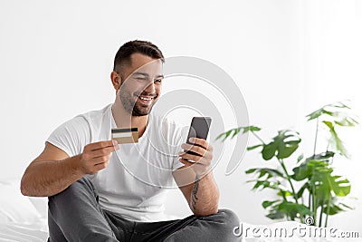 Smiling european handsome young guy in white t-shirt shopping online, orders goods on smartphone and shows credit card Stock Photo