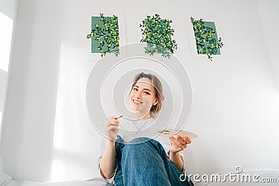 Smiling female artist sitting on bed with paintbrush, palette under her hand-drawn painting art works on the wall at Stock Photo