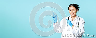 Smiling female doctor, physican in medical uniform and sterile gloves, pointing fingers left at promo, clinic logo Stock Photo