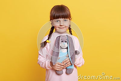 Smiling female kid with pigtails wearing pale pig shirt posing isolated over yellow background, holding soft rabbit toy in hands Stock Photo