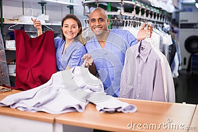 Smiling female worker of modern laundry standing with her colleague at reception counter, showing clean clothing and Stock Photo