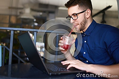 Smiling freelancer man with plastic cup of fresh beverage in hand works with laptop. Businessman in glasses drinks juice Stock Photo