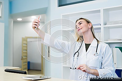 Smiling fun beautiful young doctor woman with stethoscope at hospital reception. Female doctor in medical gown doing Stock Photo