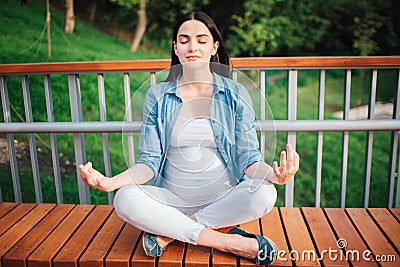 Smiling future mom expecting child sitting cross-legged and meditating with closed eyes outdoors. Prenatal Yoga. Healthy Stock Photo