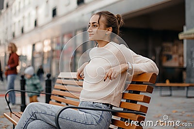 smiling girl in a knit sweater, sitting on a bench in the city in autumn. Laughing elegant woman chilling after autumn Stock Photo