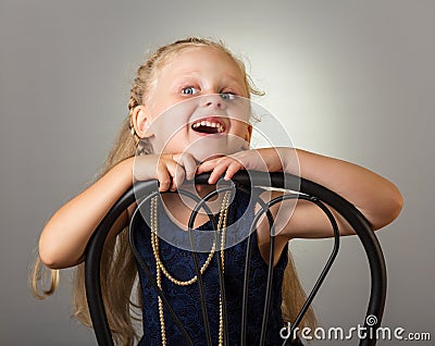 Smiling girl with long hair in dress with beads sitting on chair, on grey Stock Photo