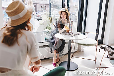 Smiling girl in long transparent dress and purple shoes sitting near window and looks at friend wears hat. Indoor Stock Photo