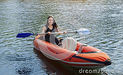 The smiling girl -the sportswoman with longdark hair in blacksportswear rows with an oar on the lake in a red inflatable canoe Stock Photo