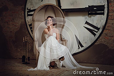 A smiling girl in a wedding dress in strange chair. The bride in a chair on the background of clocks and fireplace tool set. Stock Photo