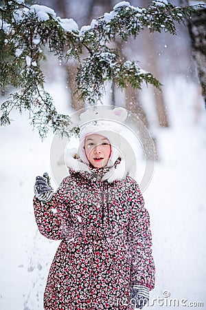 Smiling girl with white fur hat like a cat playing with snow. Winter snowy background and gteen trees Stock Photo