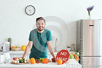 smiling handsome vegan man standing near kitchen counter with vegetables and no Stock Photo