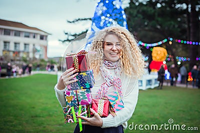 Smiling happy blonde curly woman with tower of colorful gift boxes in hands, enjoying time outdoors in decorated park with Stock Photo