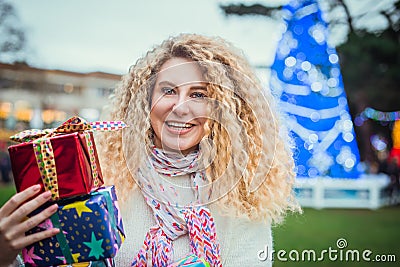 Smiling happy blonde curly woman with tower of colorful gift boxes in hands, enjoying time outdoors in decorated park with Stock Photo