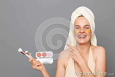 Smiling happy woman wrapped in towel standing cosmetics brushes and palette of eye shadows in hands, pointing away at Stock Photo