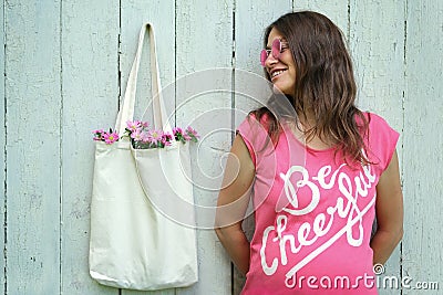 Smiling hippie woman in pink t-shirt with Be Cheerful inscription and pink round glasses standing near blank canvas eco shopping Stock Photo