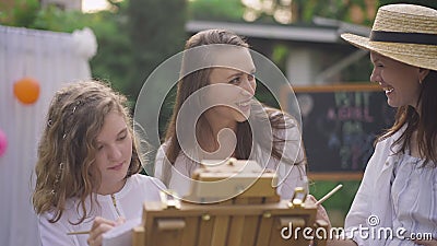 Smiling joyful girls and pregnant woman painting on easel in slow motion celebrating baby shower on backyard. Positive stock video footage Smiling joyful girls and pregnant woman painting on easel in slow motion celebrating baby shower on backyard. Positive stock video footage