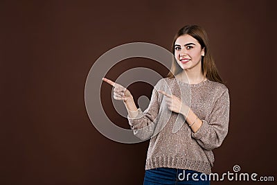 Smiling joyful woman in loose sweater posing against brown studio wall pointing at copy space for advertisment or Stock Photo