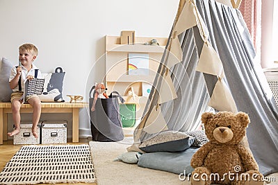 Smiling kid sitting on bench with notebook, real photo of natural playroom interior with scandinavian tent and teddy bear Stock Photo