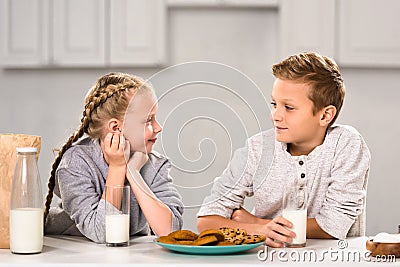 smiling kids looking at each other and sitting at table with cookies and milk Stock Photo