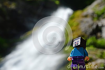 Smiling LEGO Minecraft figure of Steve in front of tall narrow mountain waterfall defocused in background. Editorial Stock Photo