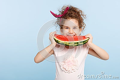 Smiling little curly girl holding with hands a large slice of watermelon, try to bite, isolated on a blue background. Stock Photo