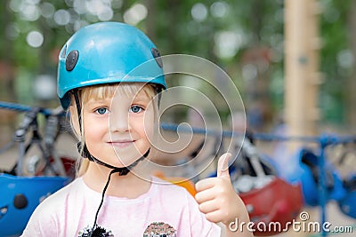 Smiling little cute blond caucasian girl in helmet showing thumbs up. Safety accessories of extreme entertainment on background. A Stock Photo