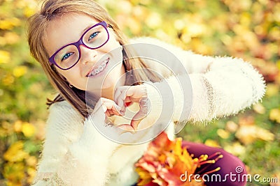 Smiling little girl with braces and glasses showing heart with hands.Autum time Stock Photo