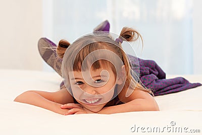 Smiling little girl lies on tummy on white bed Stock Photo