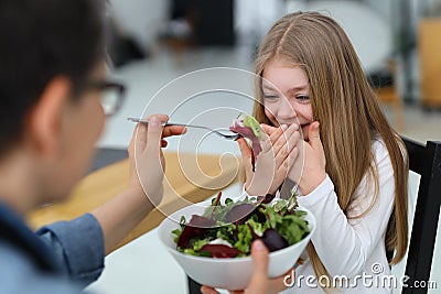 Smiling little girl refuses to eat raw salad, mother feeds her child with healthy food Stock Photo