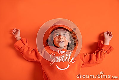 Smiling little girl 6-7 years old in cap and fashionable clothes lies on floor in the studio on orange background. Copy space Stock Photo